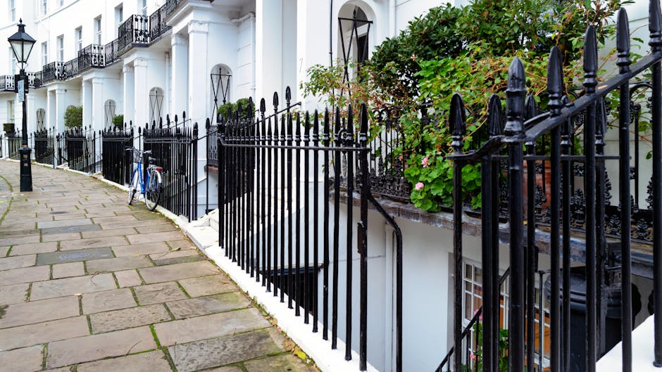 A narrow residential street with a cobblestone surface flanked by white-painted terraced houses featuring large windows and balconies. Potted plants, small trees, and shrubs are placed along the lengt