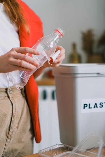 Full view of a person dressed in orange work clothing, standing on a carpeted floor within a spacious indoor environment, holding two large blue plastic trash bags filled with waste or packing materia